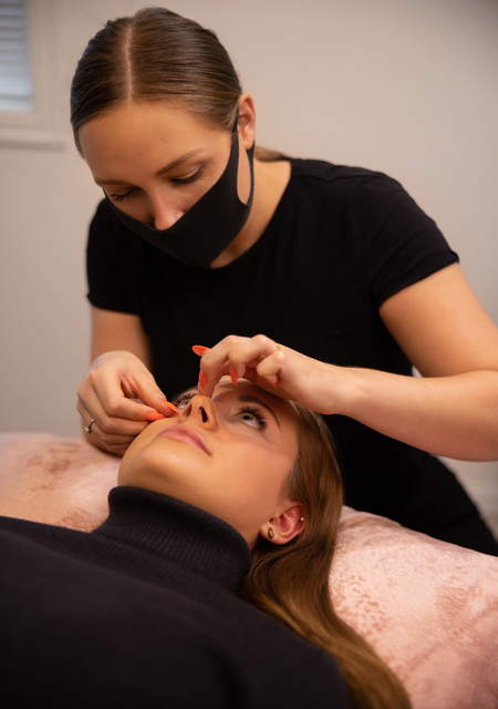 Young cosmetologist wearing protective mask treating female customer for eyelash extension at beauty spa